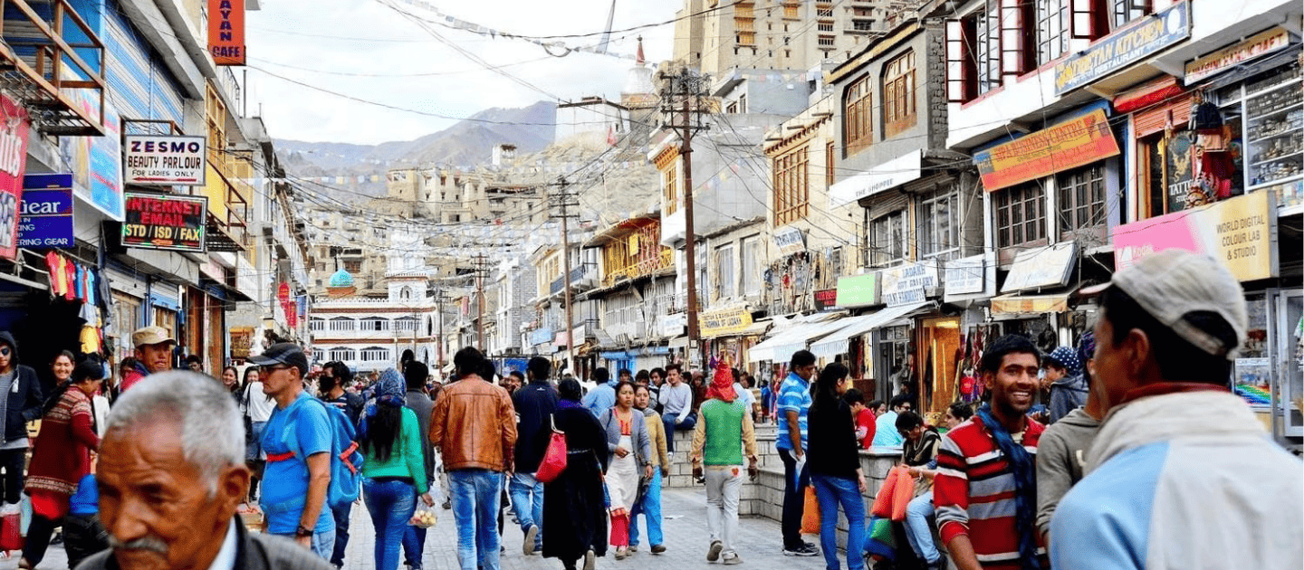 Crowded pedestrian market street in Leh Main Bazaar, Ladakh, featuring local shops, colorful signage, and tourists exploring under a cloudy mountain sky.