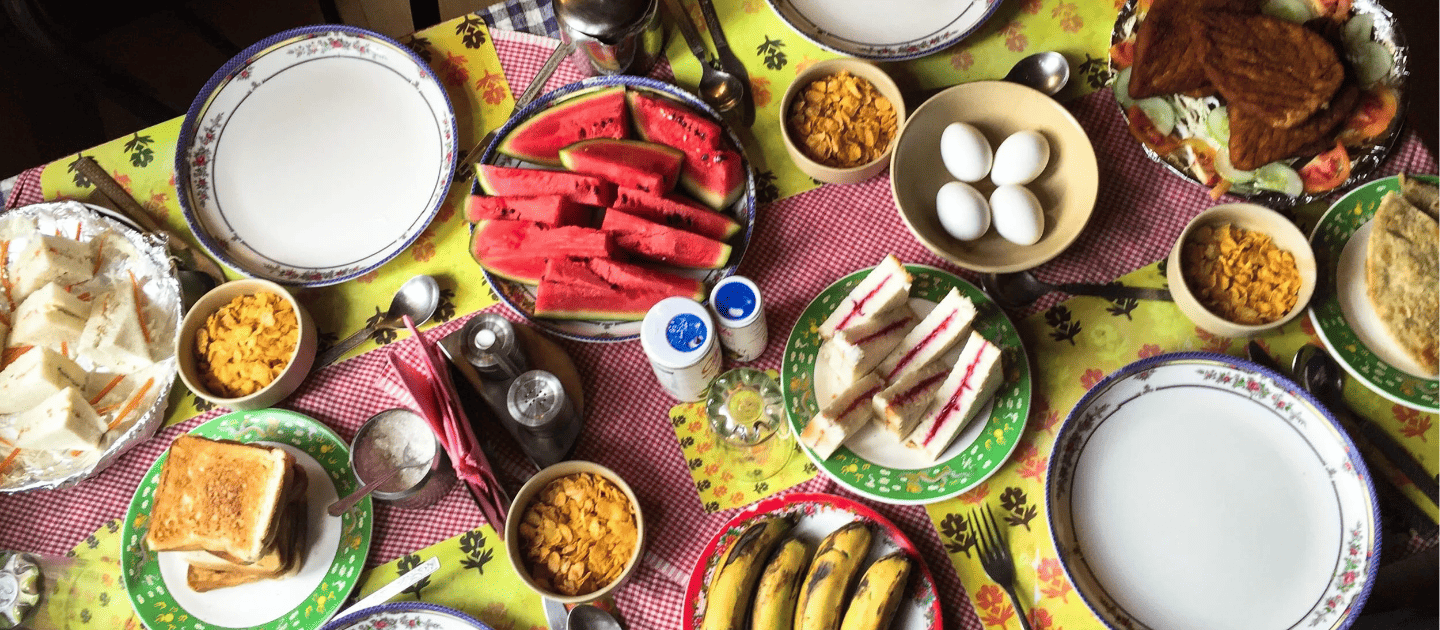 Wholesome homestay breakfast spread featuring fresh watermelon, boiled eggs, jam sandwiches, toast, and cornflakes served on a floral tablecloth