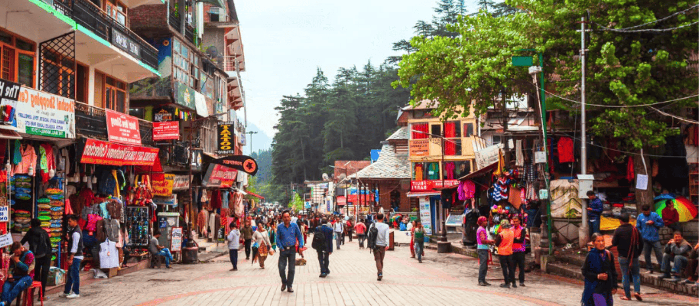 Bustling local market street in Patnitop lined with vibrant shops selling colorful winter clothes, handicrafts, and souvenirs, set against a backdrop of pine trees and mountains
